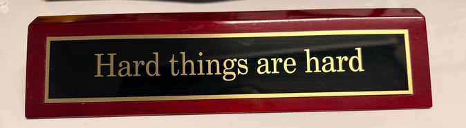 Rectangular desk plaque with a red wood frame and black center panel, featuring gold text that reads ‘Hard things are hard.’