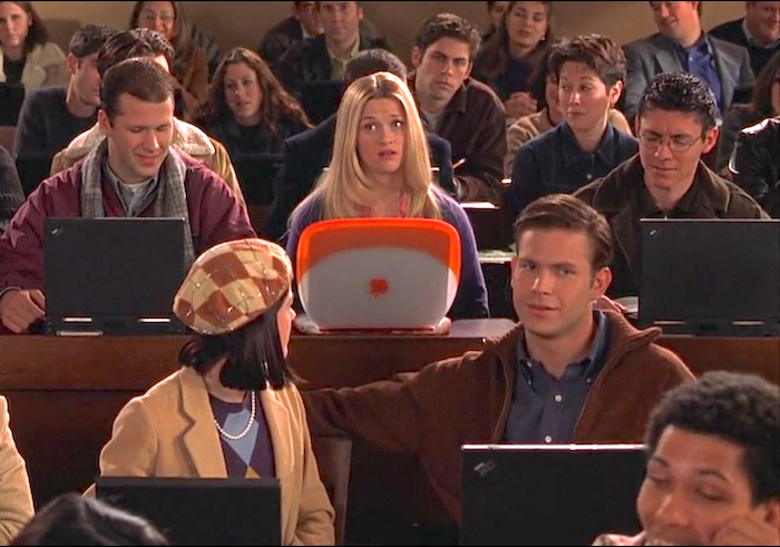 Scene from the movie Legally Blonde showing Elle Woods, played by Reese Witherspoon, sitting in a Harvard Law School lecture hall with a bright orange iBook laptop, surrounded by classmates using black laptops.