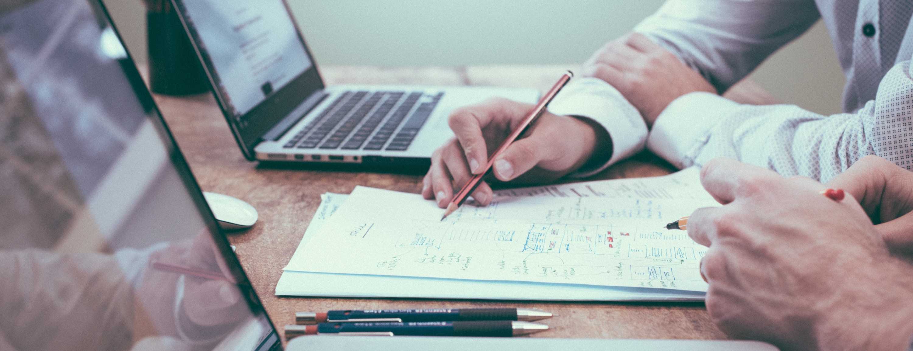 two people sitting at a table with laptops and papers.