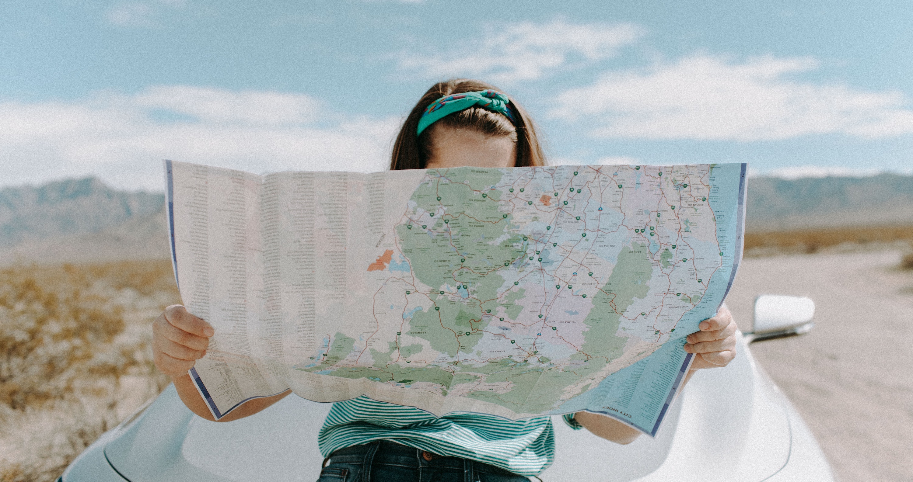 a woman leaning on a car holding a map in front of her face.