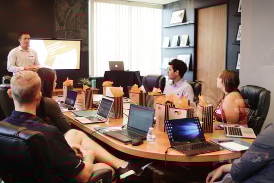 Team seated at a conference table with laptops, listening to a presenter standing beside a screen displaying a slide presentation.