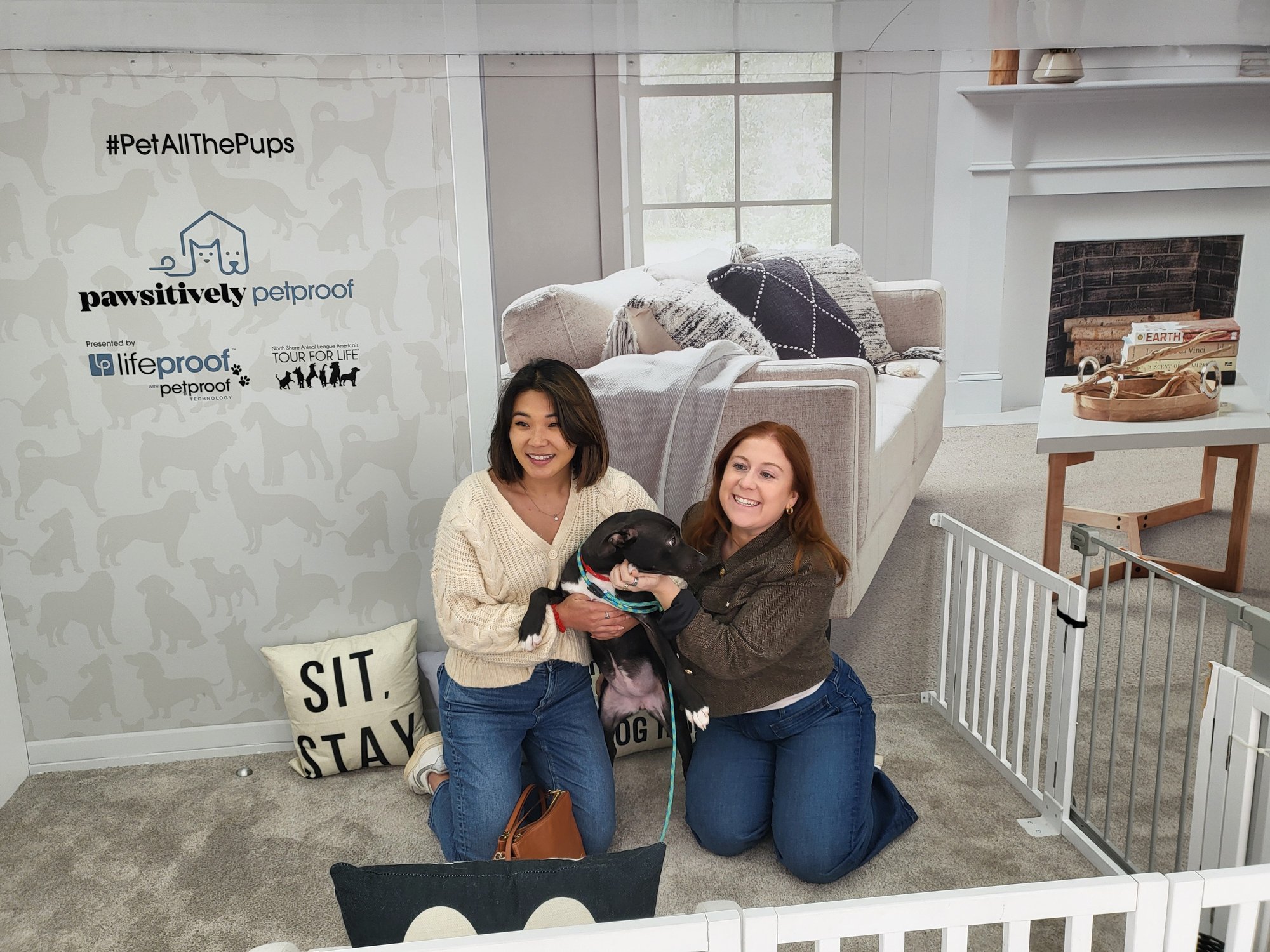 Two women kneel on a carpeted floor holding a black dog in a staged living-room setup, posing in front of a “Pawsitively Petproof” Lifeproof and Tour for Life promotional backdrop.
