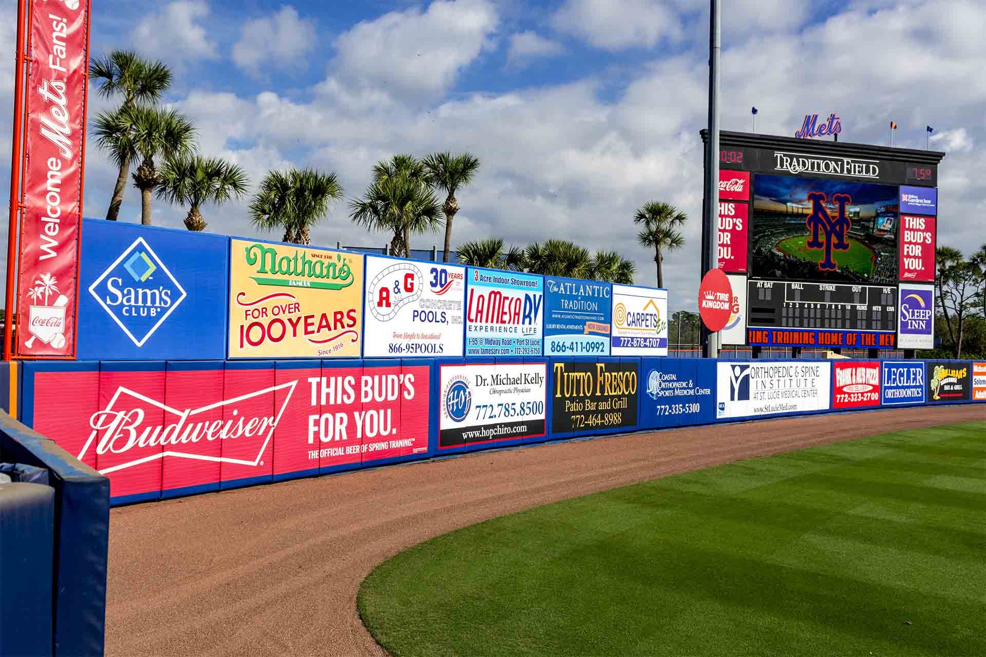 Outfield wall at Tradition Field, spring training home of the New York Mets, featuring advertisements from brands like Sam’s Club, Nathan’s Famous, Budweiser, Sleep Inn, and local businesses. A large scoreboard displays the Mets logo, with palm trees and blue skies in the background.