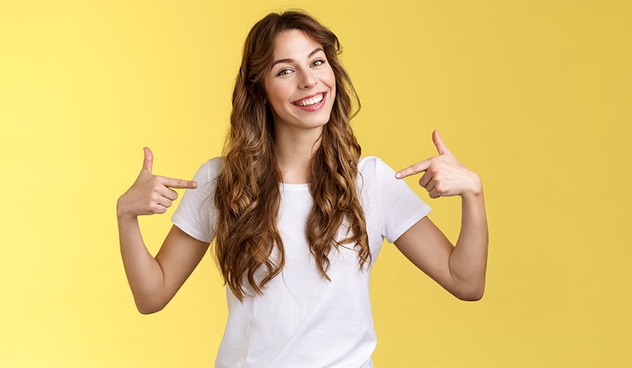 Smiling woman with long brown hair wearing a white t-shirt, standing in front of a yellow background, pointing at herself with both hands.