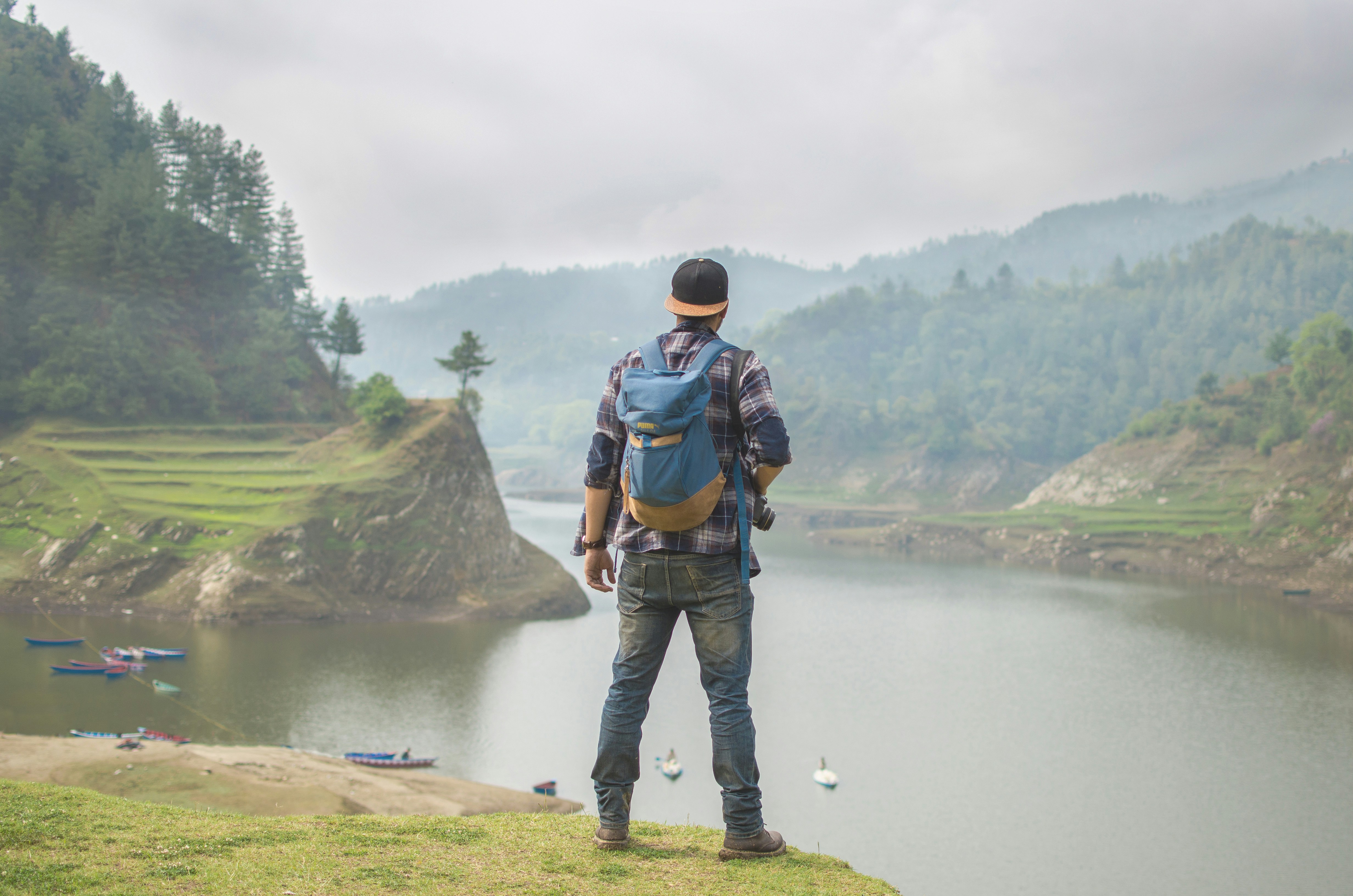 Man holding a camera while looking at a scenic lake surrounded by hills and scattered boats, under a cloudy sky.